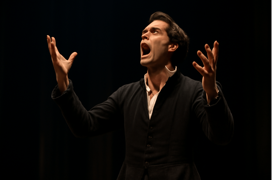 "An actor on stage performing a Shakespearean soliloquy, showing dramatic physicality. The actor’s posture should convey deep emotion, with hands raised and intense facial expression. The setting should have a dimly lit stage with spotlight on the actor, capturing the raw emotion of the soliloquy."
