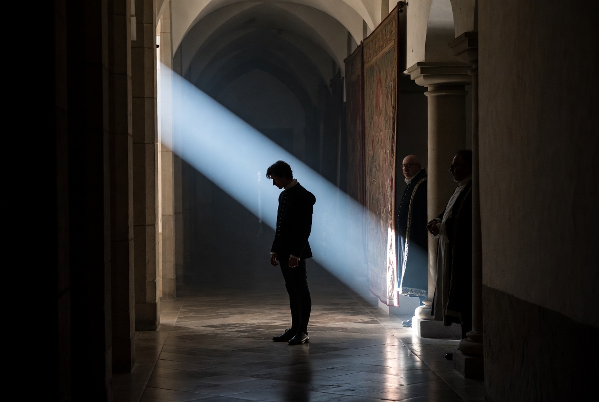 A secretive cinematic scene showing two royalty figures spying on a solitary Prince Hamlet in a dimly lit castle hallway.